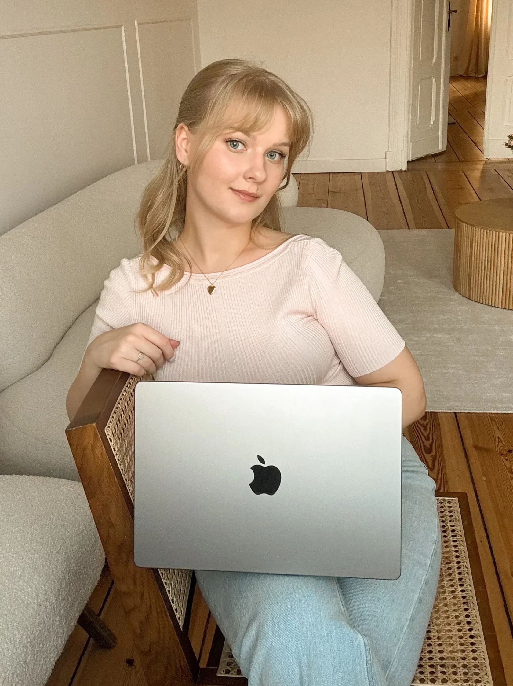 Młoda kobieta o blond włosach siedzi na drewnianym krześle i trzyma na kolanach laptopa. Ma na sobie beżowy top i niebieskie jeansy.